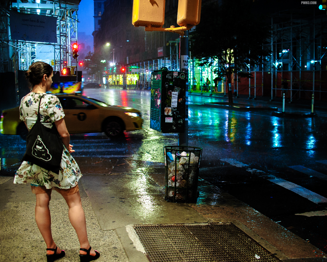 A woman waits for the rain to stop in New York City. A taxi speeds by in the distance.