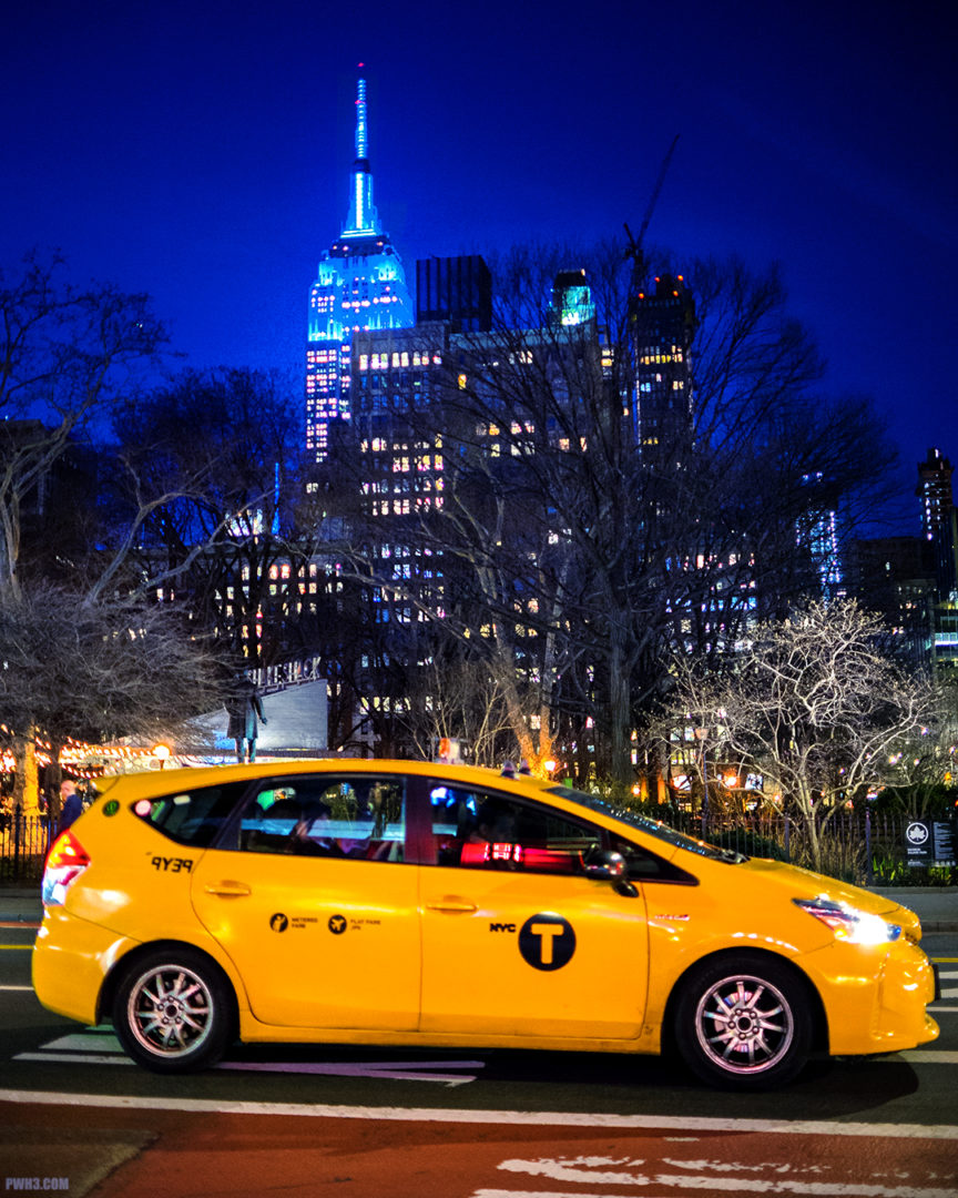 A yellow taxi waits for the light to change. Winter trees and the Empire State Building are in the distance.