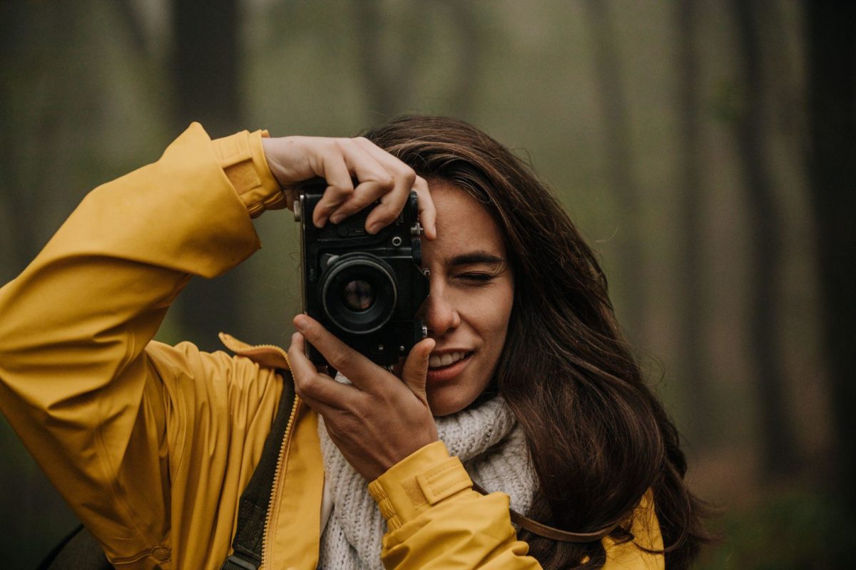 A stock photo of young woman in a forest wearing a yellow jacket, holding a camera, taking a photo as she faces us.