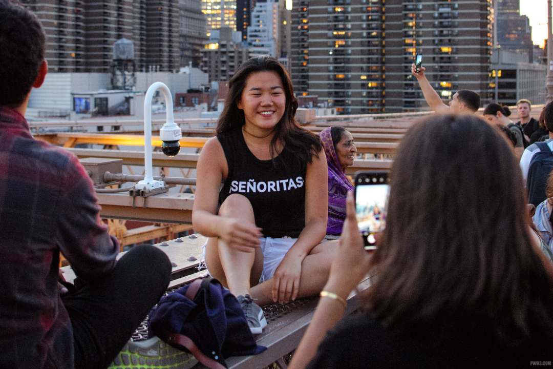 A young woman sitting on a steel girder of the Brooklyn Bridge with friends poses for a cellphone photo.